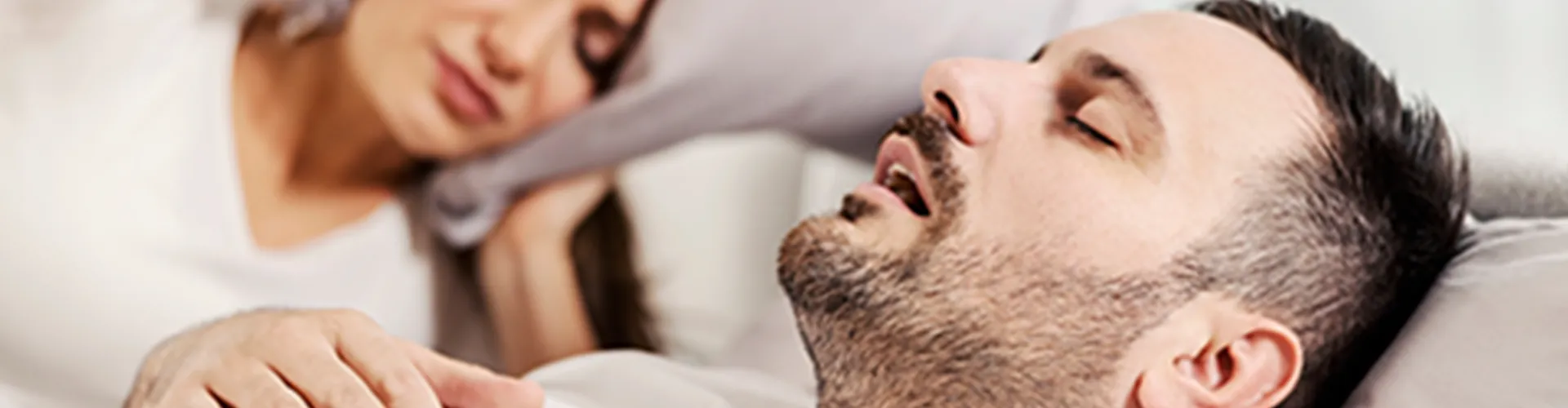 Man sleeping on his back while his wife covers her ears with a pillow.