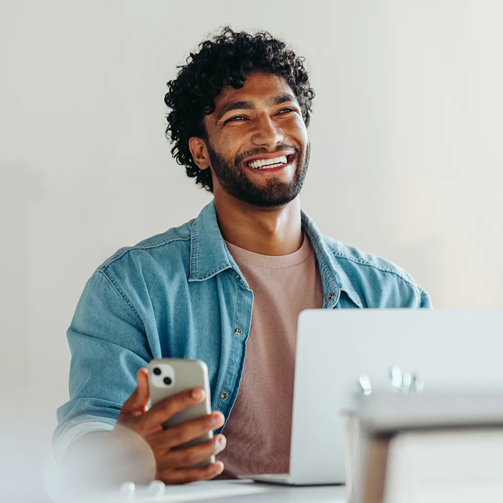 Happy man working on laptop, smiling while using smartphone.