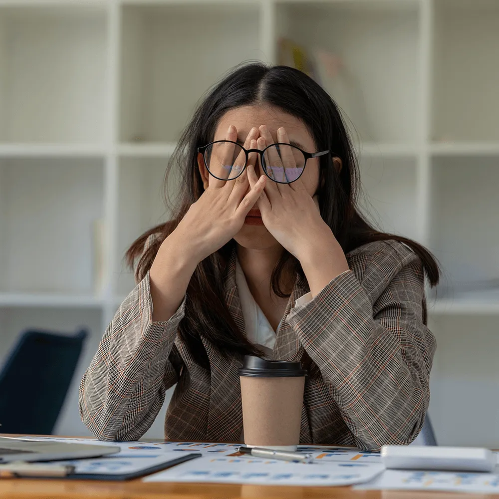 Stressed woman with glasses rubbing her eyes, sitting at a desk.