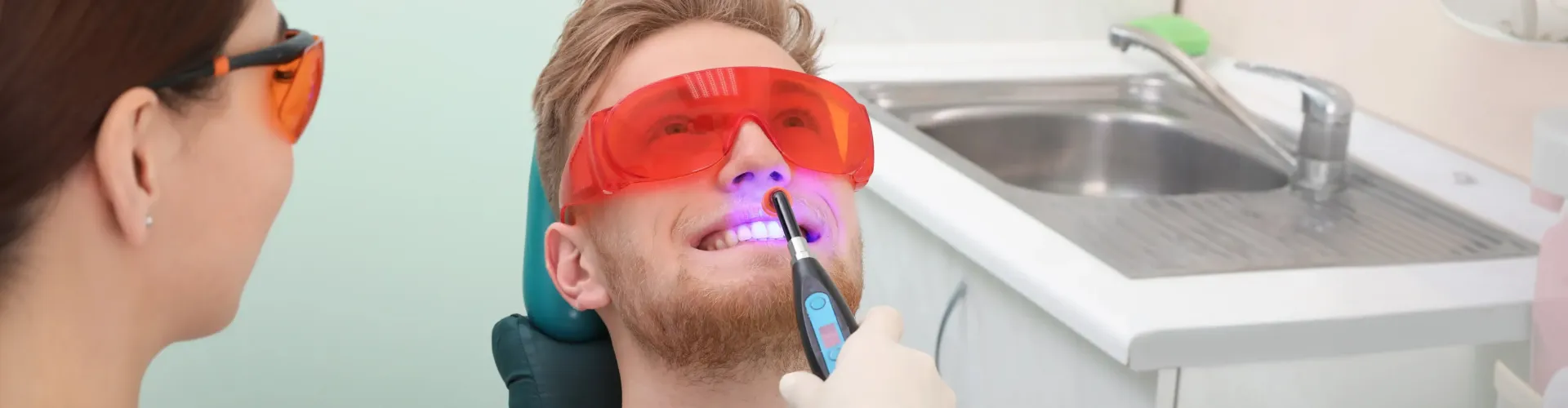Gloved hand shining blue light onto the smile of a man wearing orange eye protection.