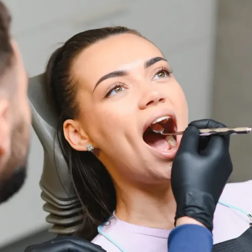 Woman receiving a dental examination while a dentist wearing black gloves uses a dental mirror to inspect her teeth in a modern dental office.