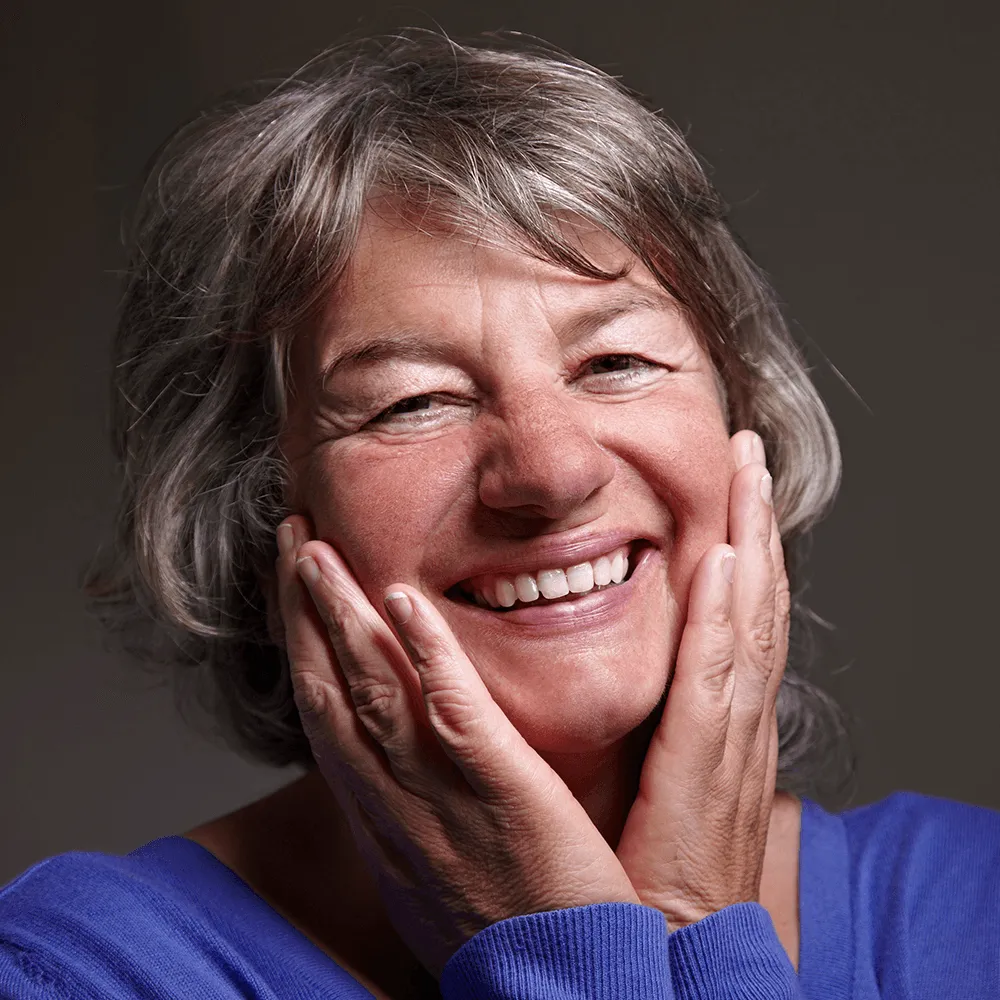Happy senior woman with gray hair, smiling broadly, hands gently touching her face.