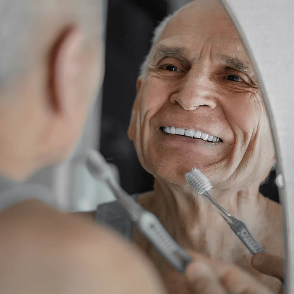 Senior man smiling while brushing his teeth in the mirror.