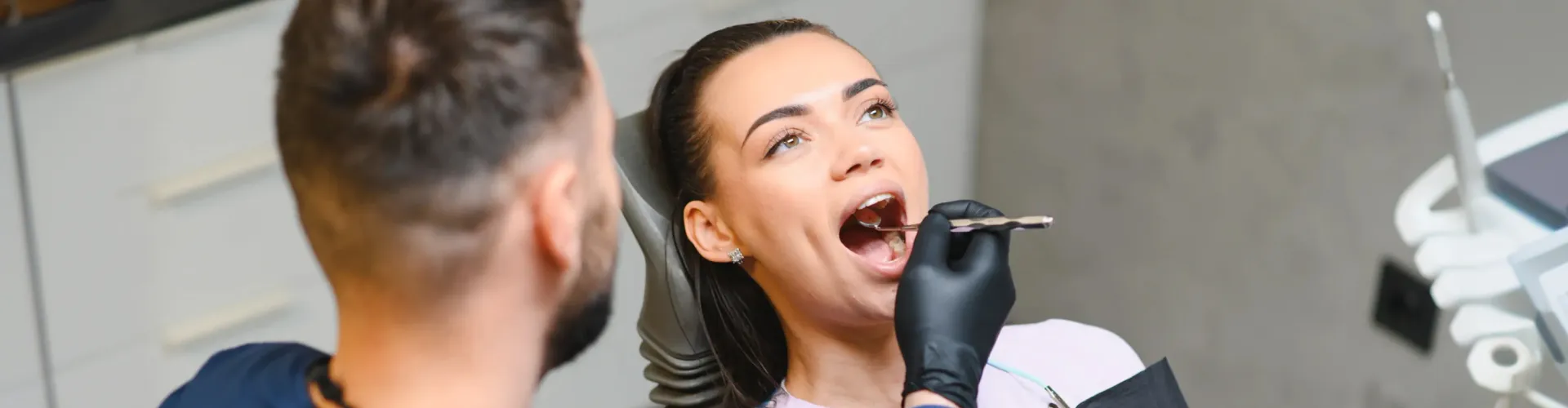 Woman receiving a dental examination while a dentist wearing black gloves uses a dental mirror to inspect her teeth in a modern dental office.