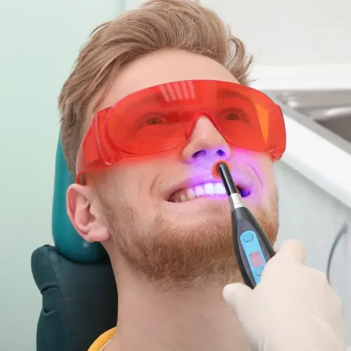 Gloved hand shining blue light onto the smile of a man wearing orange eye protection.