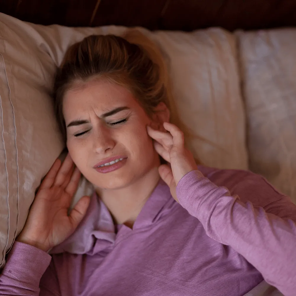 Woman in bed experiencing ear pain, holding her ears.