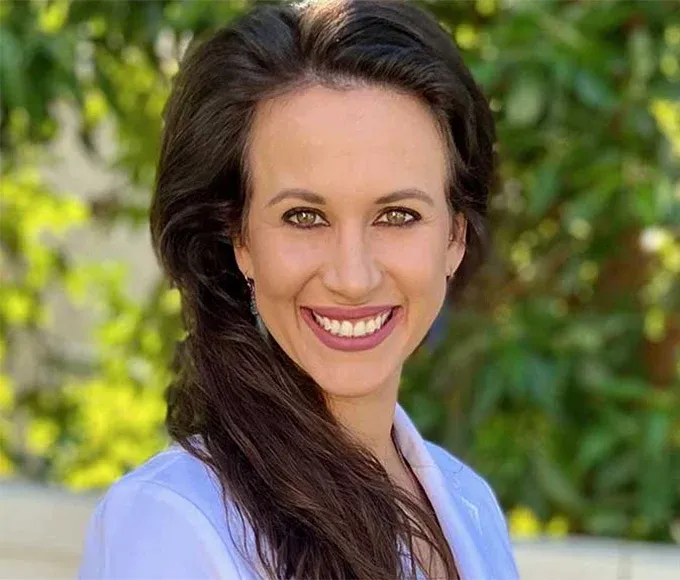 Headshot of a smiling woman with long brown hair, wearing a white blazer.