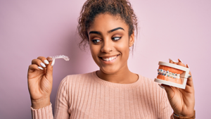 smiling woman holding clear aligner in one hand and mouth model with braces in the other